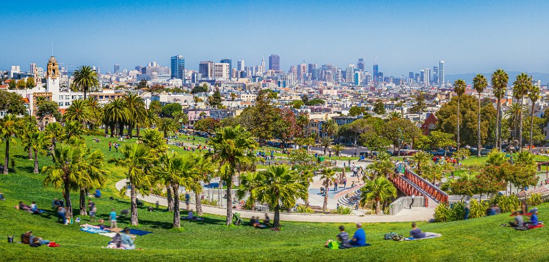 Panoramic view of local people enjoying the sunny summer weather at Mission Dolores Park on a beautiful day with clear blue sky with the skyline of San Francisco